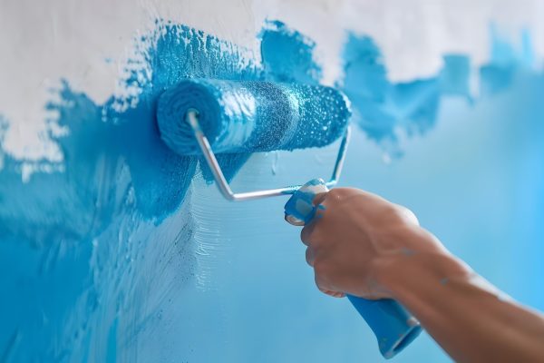 A close-up image showing a person's hand gripping a paint roller and applying blue paint to a white wall The scene depicts the process of home renovation or interior decorating,with the roller